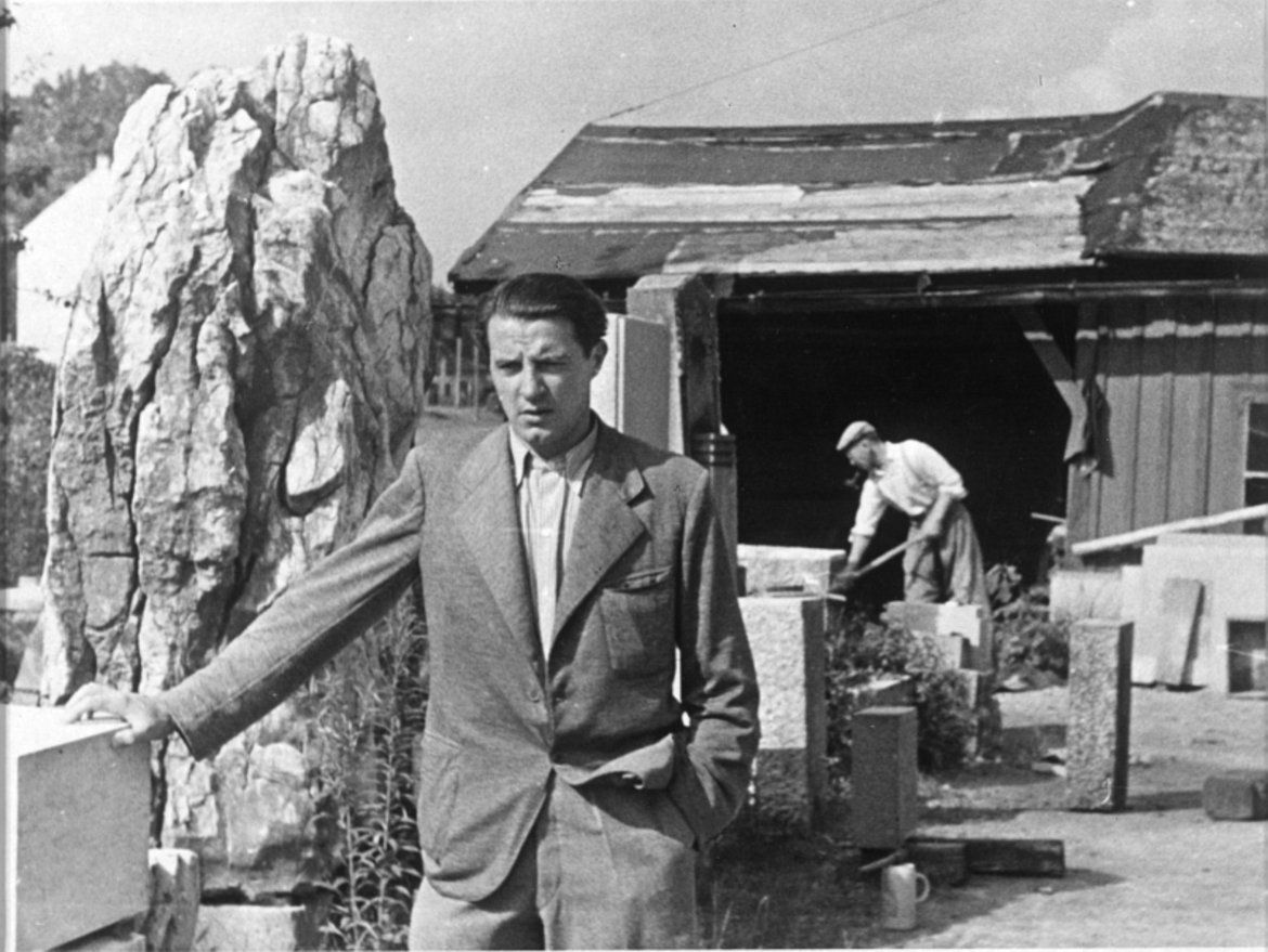 Fritz Wotruba in hellem Anzug sich lässig mit einer Hand an einem Steinquader abstützend. Im Hintergrund behauerne Steine und ein Arbeiter.