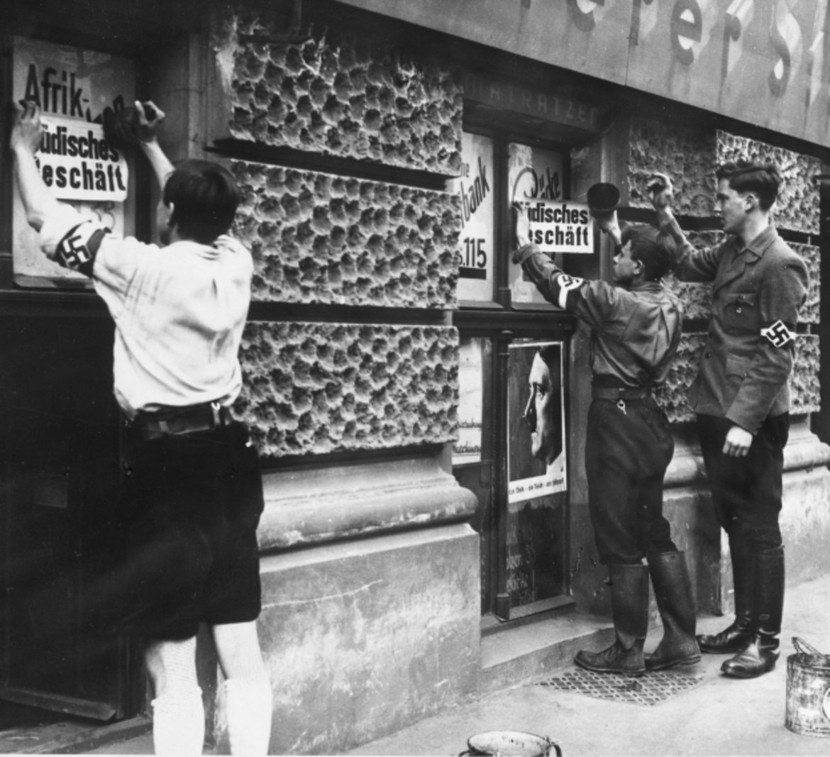 Buben, die offenbar Mittglieder der Hitler-Jugend sind, plakatieren Zetteln mit der Aufschrift "Jüdisches Geschäft" an Schaufenstern.