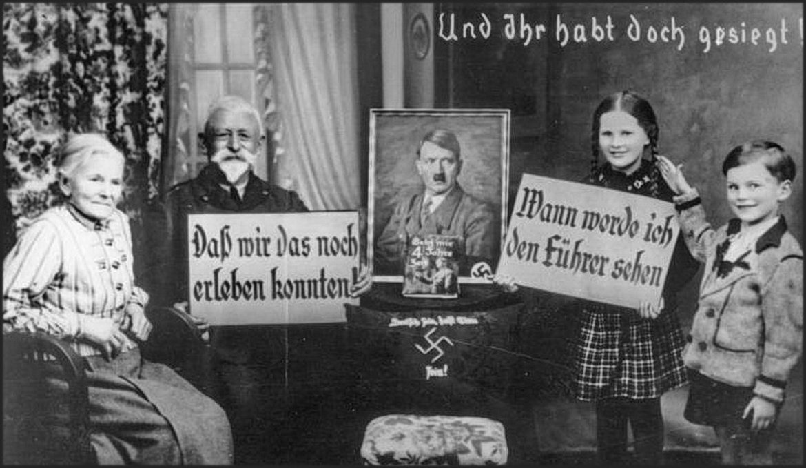 Ein altes Paar sitzt neben einem Hitlerbild. Er hält ein Schild mit der Aufschrift: "Dass wir das noch erleben konnten". Auf der anderen Seite des Hitlerbilds stehen ein Mädchen und ein Bub. Das Mädchen hält ein Schild mit der Aufschrift: " Wann werde ich den Führer sehen". 
Rechts oben auf dem Foto steht: "Und ihr habt doch gesiegt!"