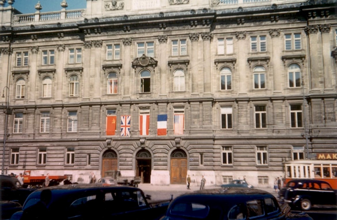 Farbfoto des Gebäudes mit den Flaggen der vier Besatzungsmächte über vier Fensterflächen. Davor einige Autos und eine Straßenbahn.
