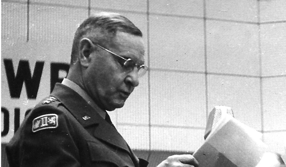 General Geoffrey Keyes vor einem Mikrofon im hellen Radiostudio in Uniform stehend.
