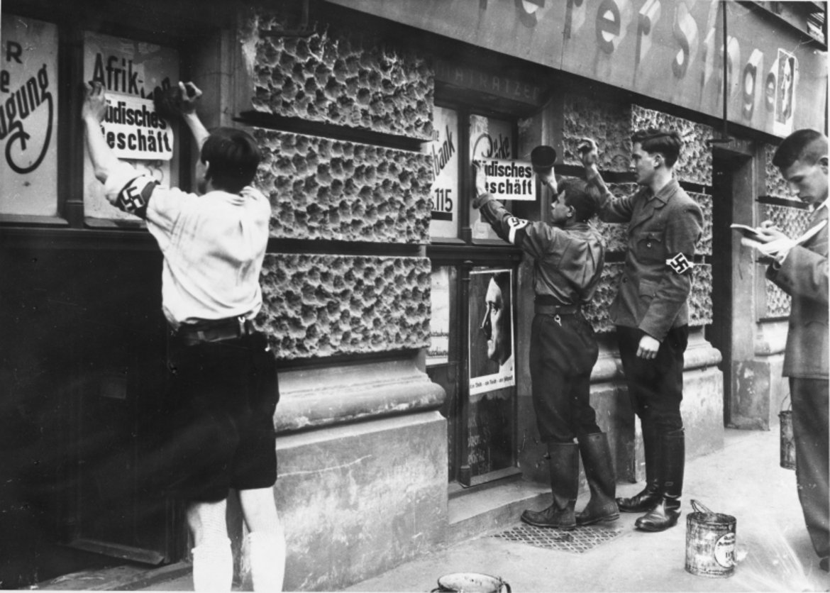 Buben, die offenbar Mittglieder der Hitler-Jugend sind, plakatieren Zetteln mit der Aufschrift "Jüdisches Geschäft" an Schaufenstern.