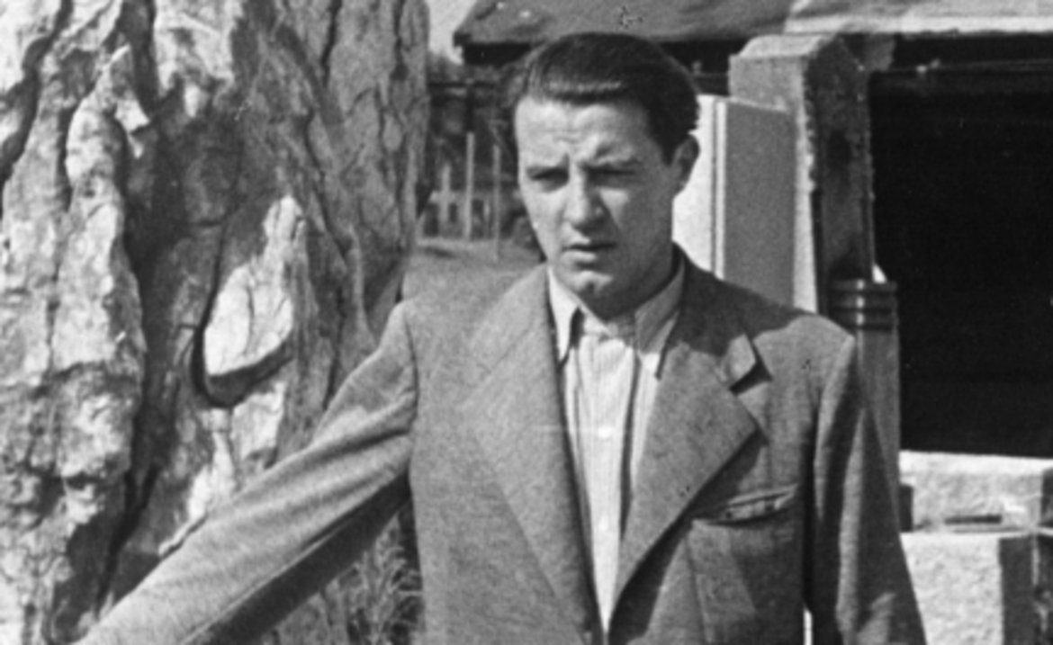 Fritz Wotruba in hellem Anzug sich lässig mit einer Hand an einem Steinquader abstützend. Im Hintergrund behauerne Steine und ein Arbeiter.