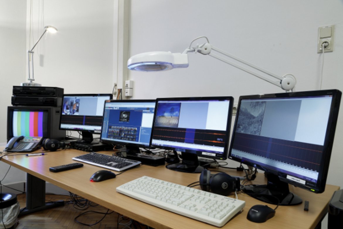 Der Arbeitsplatz in der Mediathek an dem Videos der verschiedensten Formate digitalisiert werden. Zu sehen sind fünf Monitore mit Ausschnitten aus verschiedenen Videoaufzeichnungen.