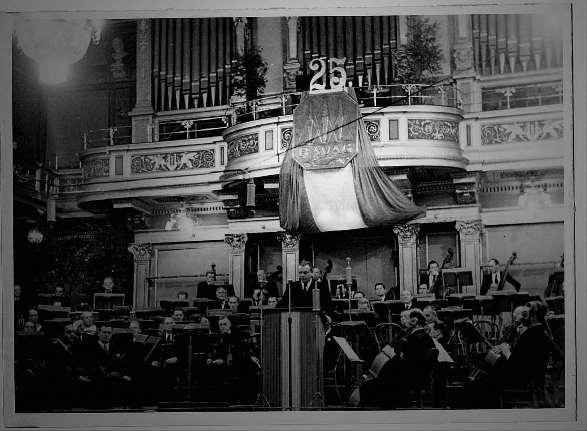 Redner am Pult vor einem sitzenden, symphonischen Orchester in einem Konzertsaal