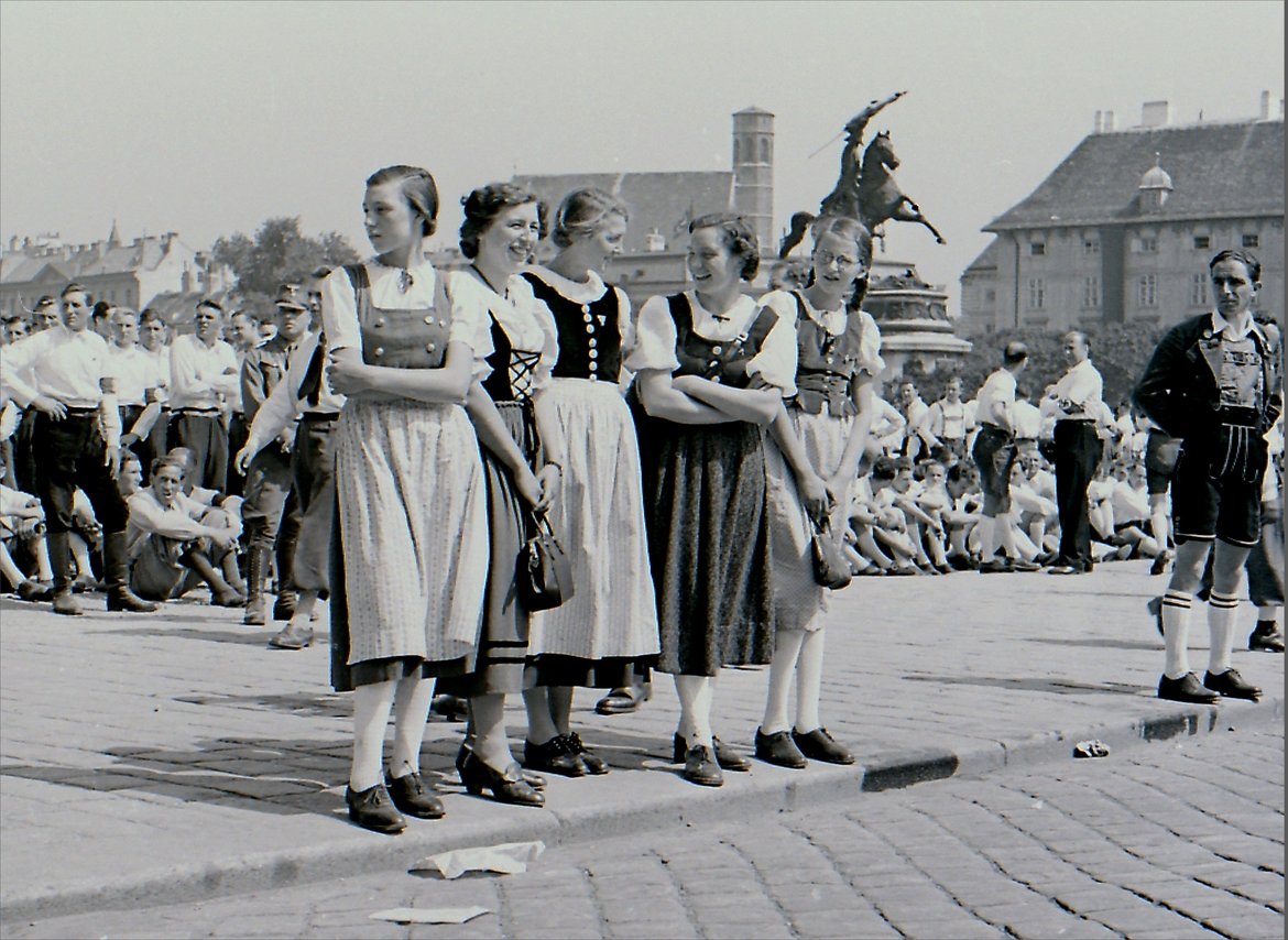 Schwarz-Weiß-Foto das viele wartende Menschen am Wiener Heldenplatz zeigt. Im Vordergrund sieht man fünf vergnügte junge Frauen in österreichischer Tracht.